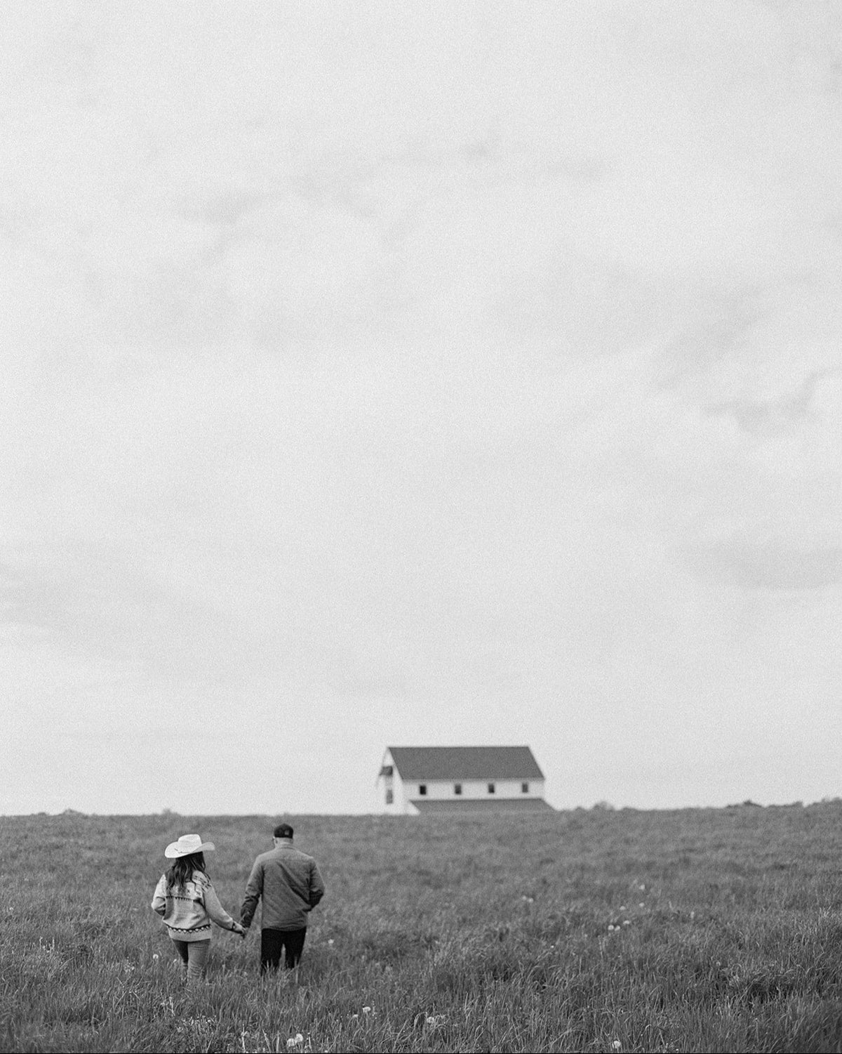 Two people walking on a grassy hill with a house in the distance under an overcast sky.