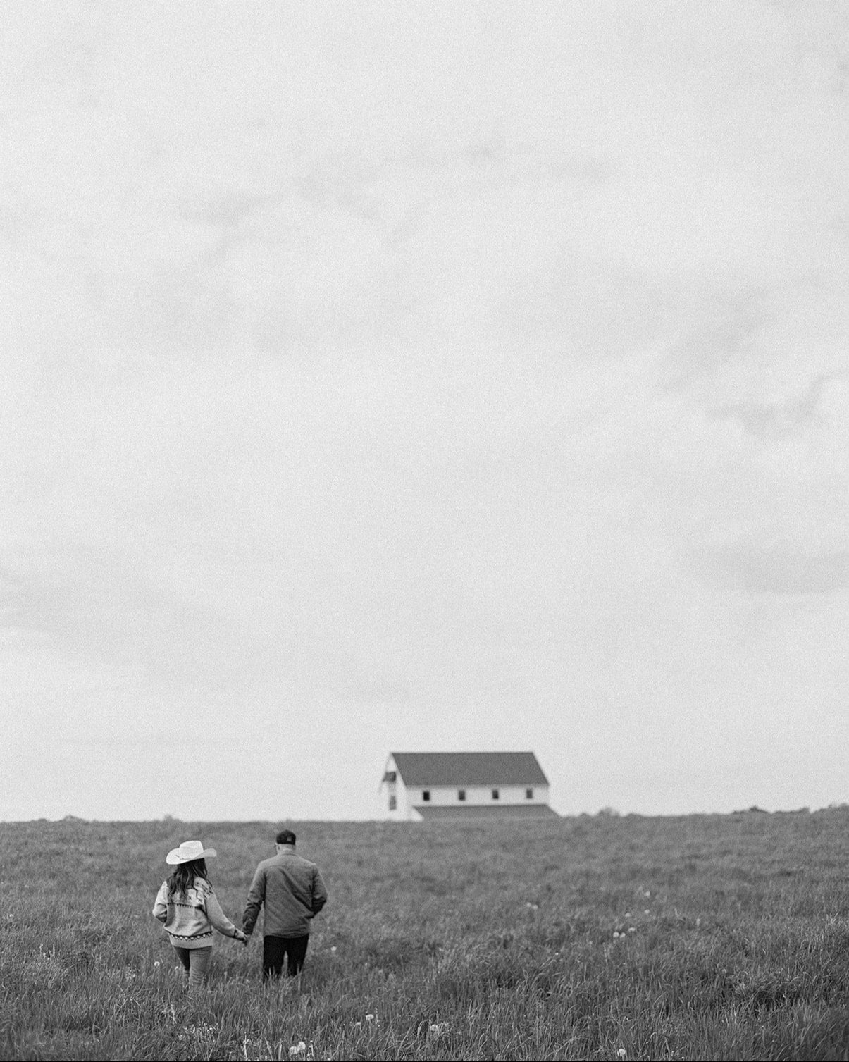 Two people walking on a grassy hill with a house in the distance under an overcast sky.