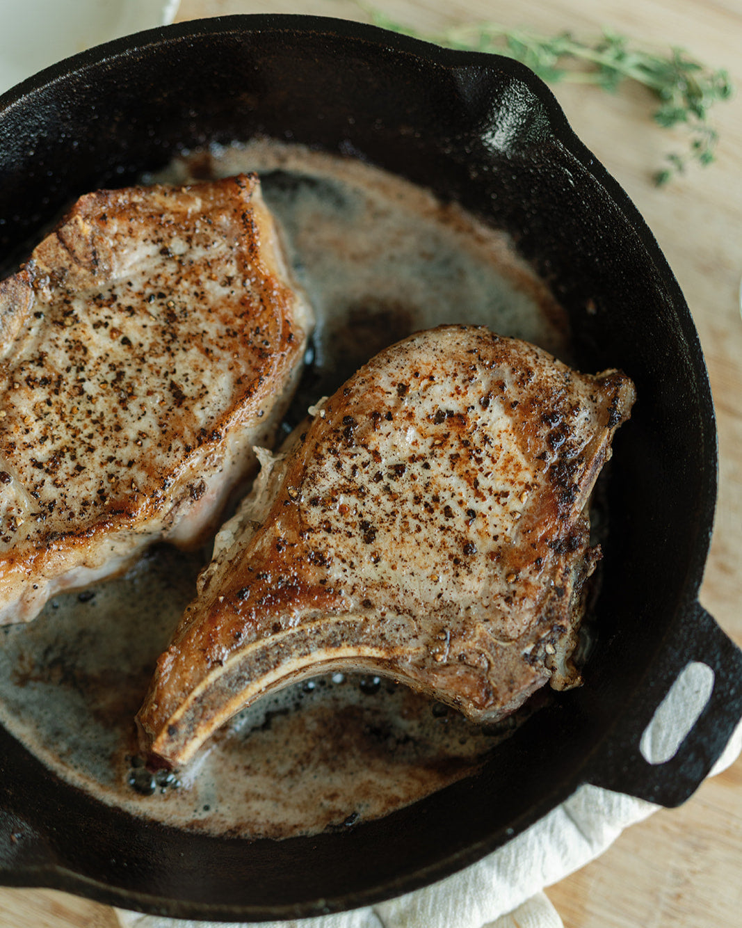 Two pork chops cooking in a black cast iron skillet on a wooden surface.