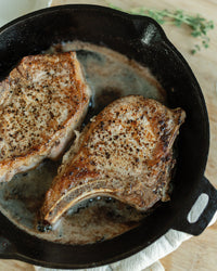 Two pork chops cooking in a black cast iron skillet on a wooden surface.